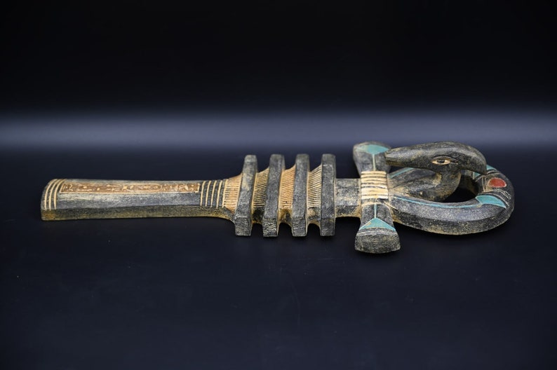 Puede incluir: Un Ankh de madera, un antiguo s&iacute;mbolo egipcio de la vida, con un acabado pintado en negro, marr&oacute;n y dorado. El Ankh es una cruz con un bucle en la parte superior y una cabeza de cobra en la parte inferior.