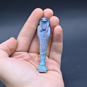 Puede incluir: Una figurita ushabti egipcia azul y morada con jerogl&iacute;ficos tallados en la superficie. La figurita tiene la forma de una persona de pie con los brazos cruzados sobre el pecho.