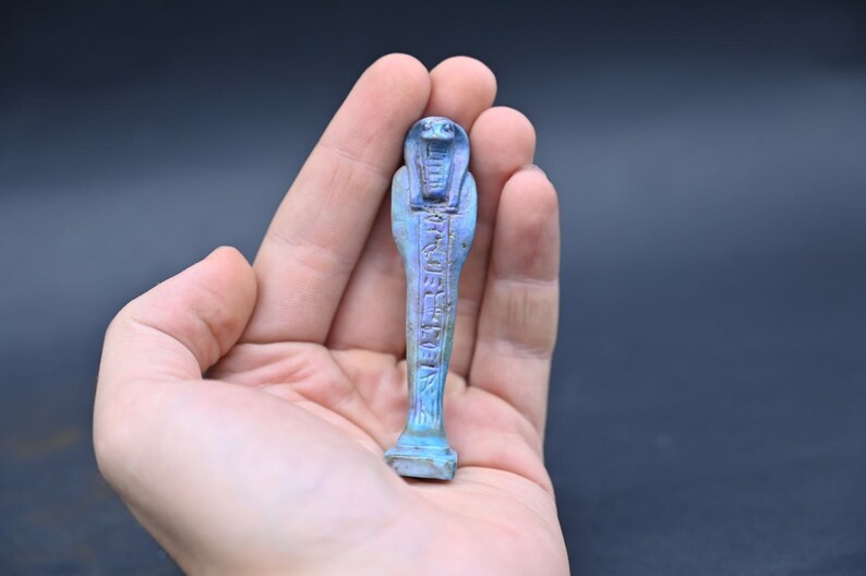Puede incluir: Una figurita ushabti egipcia azul y morada con jerogl&iacute;ficos tallados en la superficie. La figurita tiene la forma de una persona de pie con los brazos cruzados sobre el pecho.