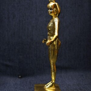 Gold leaf plated stone statue of the Egyptian goddess Neith standing with arms extended, wearing an elaborate headdress, photographed on a dark blue fabric background.