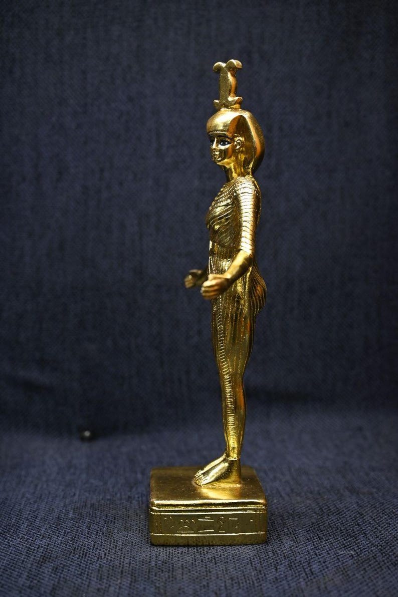 Gold leaf plated stone statue of the Egyptian goddess Neith standing with arms extended, wearing an elaborate headdress, photographed on a dark blue fabric background.