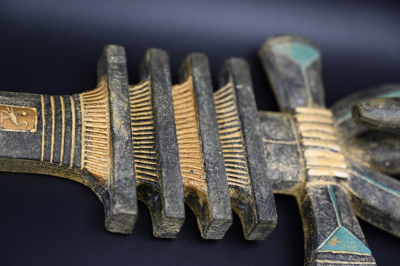 Puede incluir: Una escultura de madera negra y dorada de un ankh egipcio, s&iacute;mbolo de la vida. La escultura tiene detalles intrincados y una apariencia desgastada y envejecida.