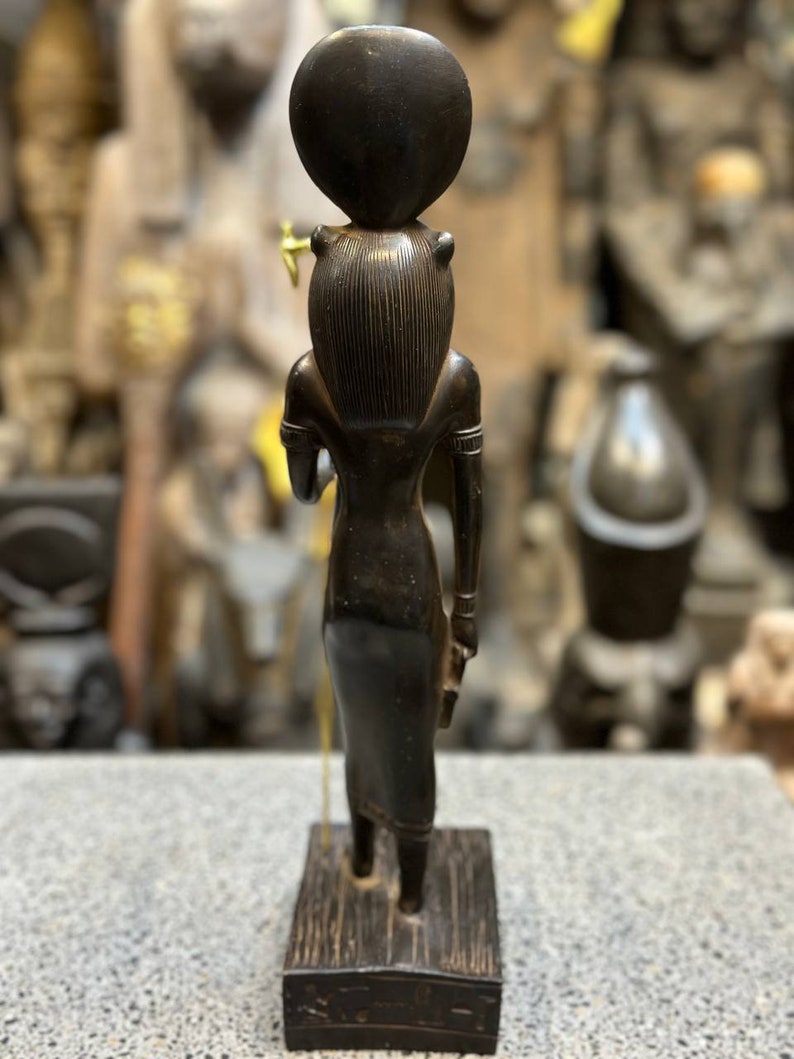 May include: Black wooden statue of an Egyptian goddess with a round headpiece and a staff in her hand. The statue is standing on a wooden base.