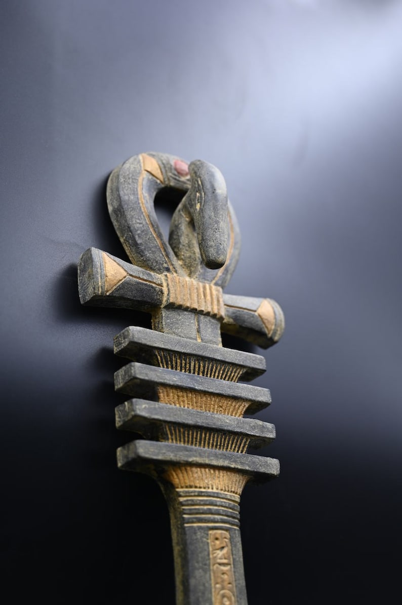 以下が含まれることがあります： グレーとゴールドの色合いの、詳細で風化したアンククロス彫刻。 上部のループは様式化された鳥の頭部が特徴です。 十字バーとベースには幾何学模様があります。 彫刻は暗い背景に配置されています。