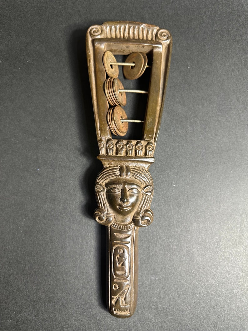 May include: A wooden sistrum with a carved female head and a series of metal discs that rattle when shaken. The sistrum is a musical instrument used in ancient Egypt.