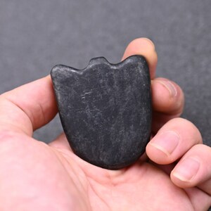 May include: A black, smooth, heart-shaped stone with a slightly raised edge. The stone is held in a hand.