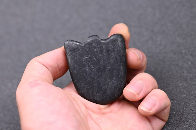 May include: A black, smooth, heart-shaped stone with a slightly raised edge. The stone is held in a hand.