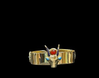 May include: A gold ring with an Egyptian bull head design. The ring has a red stone, blue accents, and a wide band. The ring is set against a black background. The ring's band is approximately 2 cm wide.