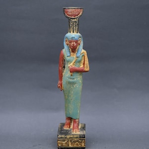 May include: A small, painted figurine of an Egyptian goddess standing on a square base. The goddess is wearing a blue headdress and a green robe. She is holding a staff in her right hand.