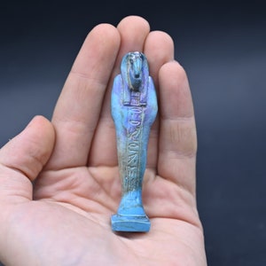 Puede incluir: Una figurita ushabti egipcia azul y morada con jerogl&iacute;ficos tallados en la parte delantera. La figurita est&aacute; de pie sobre una peque&ntilde;a base.