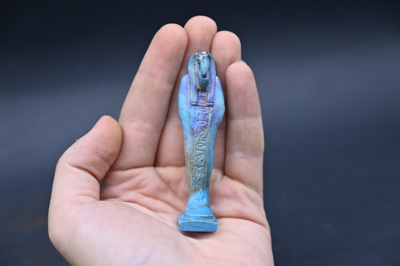 Puede incluir: Una figurita ushabti egipcia azul y morada con jerogl&iacute;ficos tallados en la parte delantera. La figurita est&aacute; de pie sobre una peque&ntilde;a base.