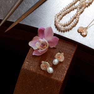 May include: A still-life image featuring jewelry and floral elements. A pair of pearl earrings and a pendant necklace with a flower design are displayed alongside a strand of pearls. A pink orchid flower rests on a wooden surface.