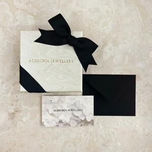 May include: A white gift box with a black ribbon and bow. The box is tied with a black ribbon and bow. The box has the text "ALEGORIA JEWELLERY" printed on the front. A white card with the text "ALEGORIA JEWELLERY" printed on it is next to the box.