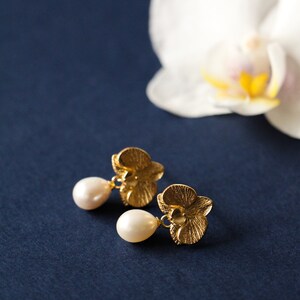 May include: A pair of gold orchid flower earrings with pearl drops. The earrings are on a dark blue background.