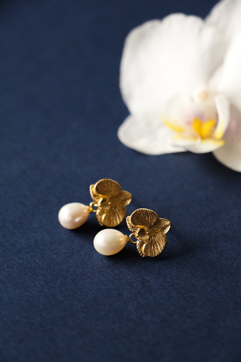 May include: A pair of gold orchid flower earrings with pearl drops. The earrings are on a dark blue background.