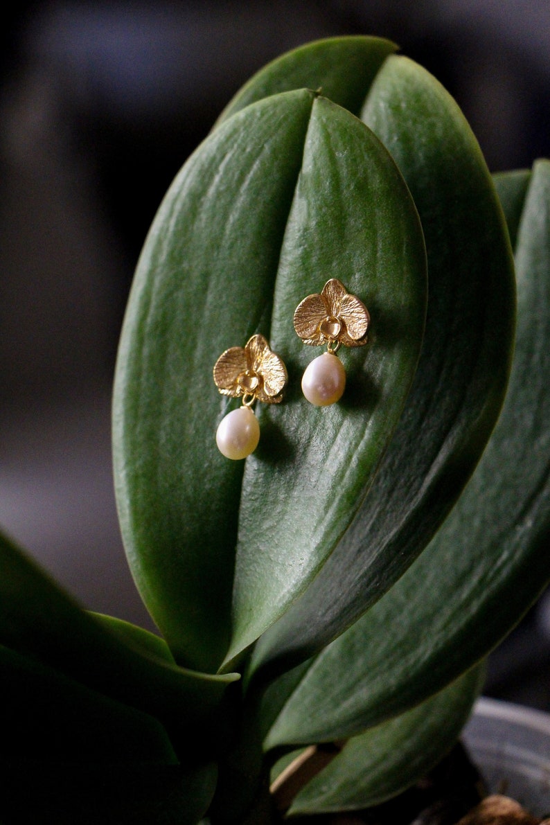 May include: A pair of gold orchid earrings with pearl drops. The earrings are displayed on a green leaf.