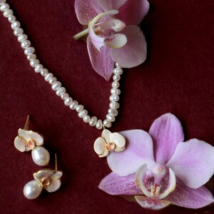May include: A pearl necklace and matching earrings with orchid pendants are displayed on a burgundy background, alongside two pink orchid flowers. The jewelry features small, round pearls and gold-toned metal accents.
