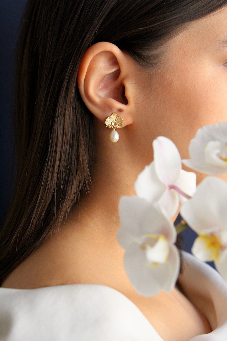 May include: Gold orchid flower earrings with a pearl drop. The earrings are worn by a person with long brown hair and a white shirt.
