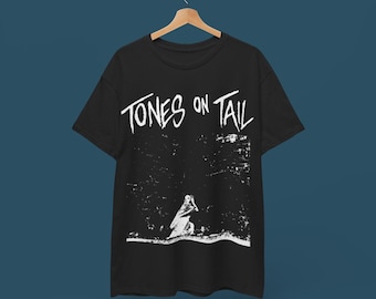 May include: Black t-shirt featuring the white text "TONES ON TAIL" and a white graphic of a kneeling figure. The t-shirt is displayed on a wooden hanger.