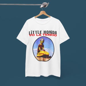 May include: White t-shirt with a graphic of a person riding a yellow snowmobile. The text "LITTLE HONDA YO LA TENGO" is printed on the shirt.