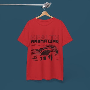 May include: A red t-shirt with the words "HEALTH ARENA WAR" printed in black block letters above a black graphic of a vehicle. The t-shirt is hanging on a wooden hanger.