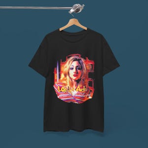 May include: A black short-sleeve t-shirt featuring a graphic print of a person with blonde hair wearing a striped shirt. The text "Lords of Acid" and "Voodoo-U" is printed in yellow and white below the portrait, set against a red abstract background.