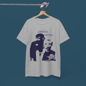 May include: A gray t-shirt featuring a navy blue graphic of two figures and the text "CRYSTAL CASTLES" in a sans-serif font. The shirt is hanging on a wooden hanger against a blue background.