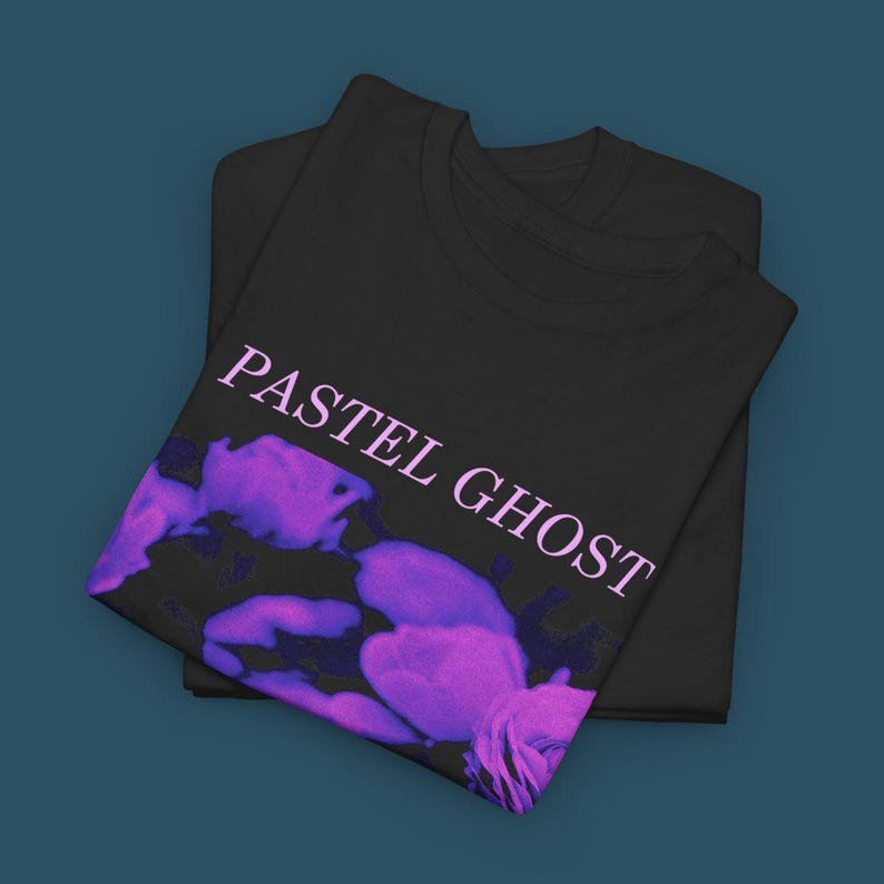 May include: Two black t-shirts are stacked on a dark blue surface. The top shirt features the text "PASTEL GHOST" in a light purple color, along with a purple and blue abstract design.