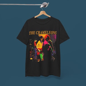 May include: A black short-sleeve t-shirt featuring a graphic print of a band performing on stage. The text THE CHAMELEONS is printed in yellow above the colorful illustration of musicians playing instruments under stage lighting.