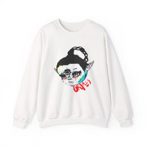 May include: White crewneck sweatshirt featuring a colorful illustration of a three-eyed face with elf-like ears. The word "GRIMES" is written in red script below the face. The design is centered on the front of the sweatshirt.