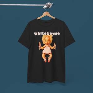 May include: Black t-shirt with the word "whitehouse" in white and red. The shirt displays a cartoon doll holding bottles. The doll wears a white dress with red trim and red shoes. The t-shirt is hanging on a wooden hanger.