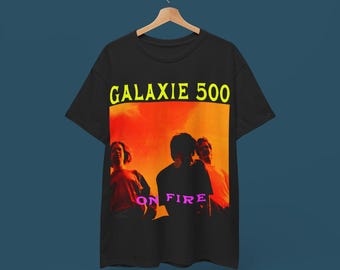 Puede incluir: Camiseta negra con un gráfico de la banda Galaxie 500. El gráfico es una foto de los miembros de la banda frente a una puesta de sol con el texto "On Fire" en rosa.