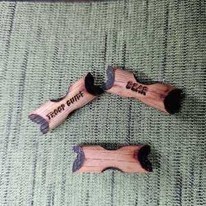 May include: Three wooden bear-shaped cutouts with the words "TROOP GUIDE" and "BEAR" etched into them. The cutouts are light brown with black accents and are arranged on a green textured surface.