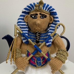 May include: A knitted teapot cozy in the shape of an Egyptian pharaoh. The cozy is brown with blue and gold accents. The pharaoh is wearing a blue and white striped headdress, a gold collar, and a gold belt. The cozy is holding two golden staffs.