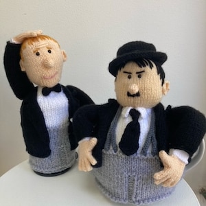 May include: Two knitted tea cozy figures, one with a black suit and a white bow tie, the other with a black suit, a gray vest, and a black tie. The figures are in a classic comedy duo style.