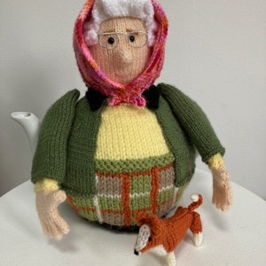 Puede incluir: Una funda de tetera de punto con forma de mujer que lleva una bufanda rosa y roja, un cárdigan verde y una falda a cuadros. Lleva un pequeño zorro de punto.