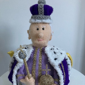 May include: A knitted teapot cover depicting a royal figure. The cover features a purple and silver crown, a purple and gold robe, and holds a gold scepter and orb. The teapot spout is yellow.