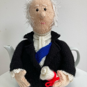 May include: Knitting pattern for a tea cosy shaped like a person wearing a black suit, white shirt, blue sash, and holding a scroll. The person has white hair and a pink face.