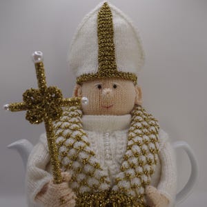 Könnte beinhalten: Handgestrickte Teewärmer in Form eines Papstes, komplett mit Mitra, Kreuz und goldenen Akzenten. Der Teewärmer ist weiß mit goldenen Verzierungen und einem dekorativen goldenen Kreuz. Er ist für eine Teekanne konzipiert.