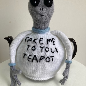 Puede incluir: Una funda de tetera de punto gris en forma de extraterrestre con ojos negros y el texto "Take me to your teapot" bordado en negro.