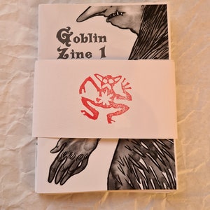 May include: A black and white illustration of a goblin on the cover of a zine titled "Goblin Zine 1". The zine is wrapped in a white paper band with a red goblin stamp.