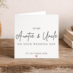 May include: A square white greeting card with black text that reads TO MY Auntie & Uncle ON YOUR WEDDING DAY, followed by the date 1ST OCTOBER 2026 and a small pink heart icon centered at the bottom of the card face on a textured paper surface.