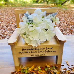 May include: A small wooden chair with a white flower arrangement on top. The chair has a message engraved on the front that reads "Remembering our loved ones who have gone before us. We miss you x".
