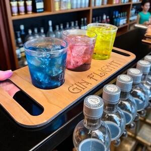 May include: A wooden serving tray with the words "GIN FLIGHT" engraved on it, holding three clear plastic cups filled with colourful cocktails and ice. The drinks are blue, pink, and yellow. Several gin bottles are in the foreground.