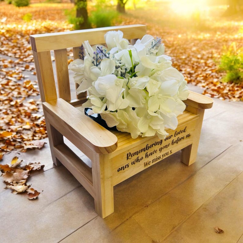 May include: A small wooden bench with a white flower arrangement on top. The bench has a personalized inscription that reads "Remembering our loved ones who have gone before us. We miss you x".