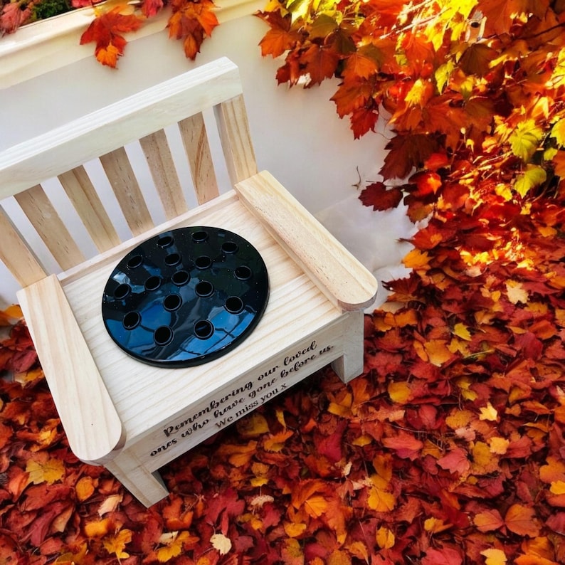 May include: A wooden memorial bench with a black metal insert for flowers. The bench has the inscription "Remembering our loved ones who have gone before us. We miss you X". The bench is sitting on a bed of red and orange leaves.