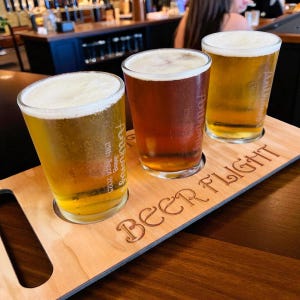 May include: A wooden beer flight tray with three glasses of beer. The beers vary in colour from light gold to amber, each topped with a foamy head. The tray is engraved with the words "BEER FLIGHT". The glasses have text on them.