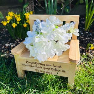 May include: A small wooden chair with a white flower arrangement on top. The chair has a message engraved on the front that reads "Remembering our loved ones who have gone before us. We miss you x".