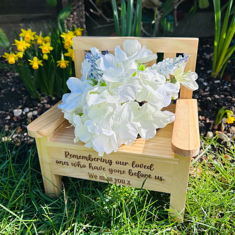 May include: A small wooden chair with a white flower arrangement on top. The chair has a message engraved on the front that reads "Remembering our loved ones who have gone before us. We miss you x".