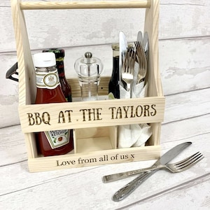 May include: Wooden crate with a handle, personalised with the text "BBQ at the Taylors" and "Love from all of us x". The crate contains bottles of ketchup, sauce, and oil, a salt and pepper shaker, and cutlery.